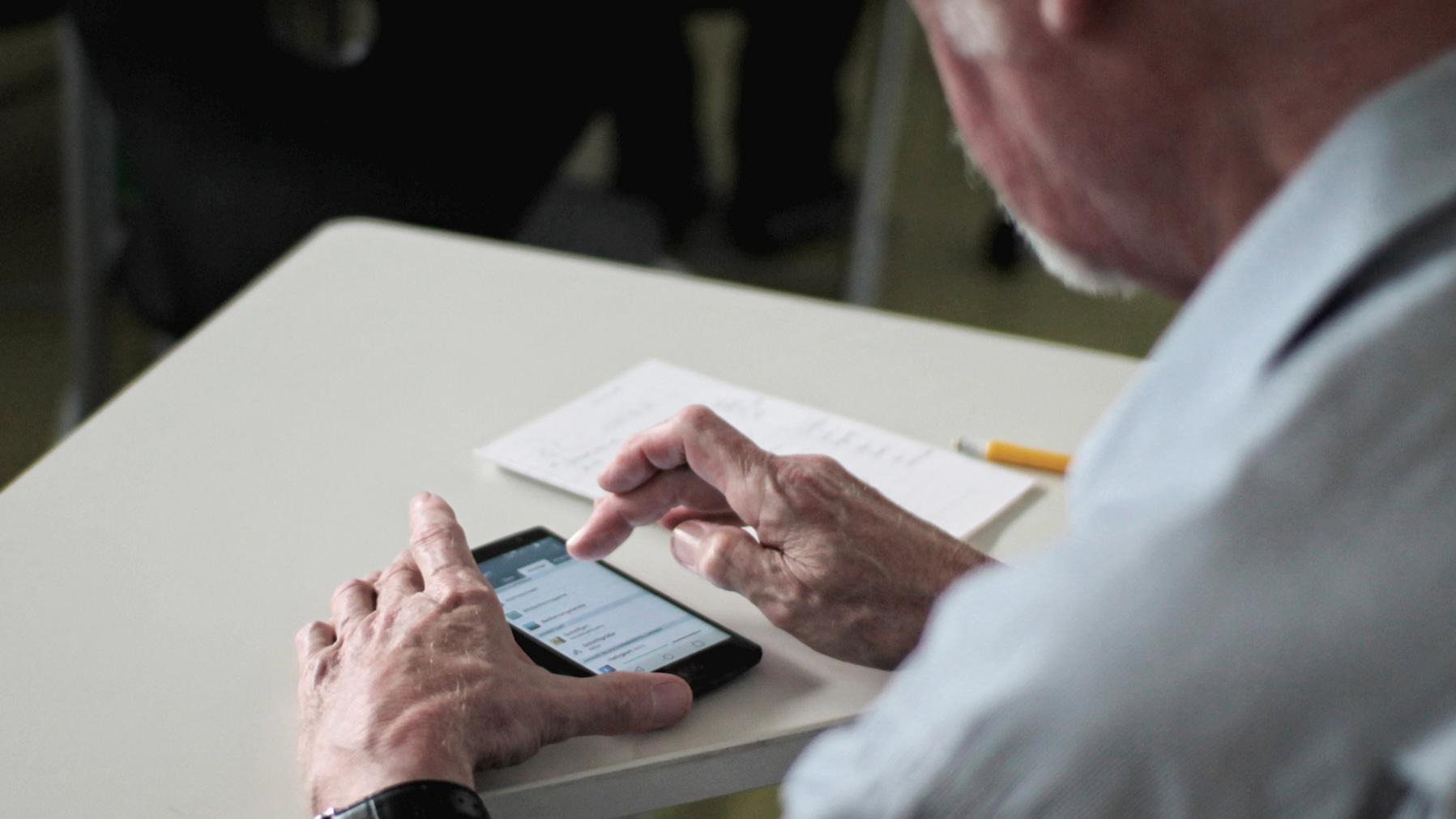 Älterer Mann, der an einem weißen Tisch sitzt und mit beiden Händen ein Smartphone bedient Foto